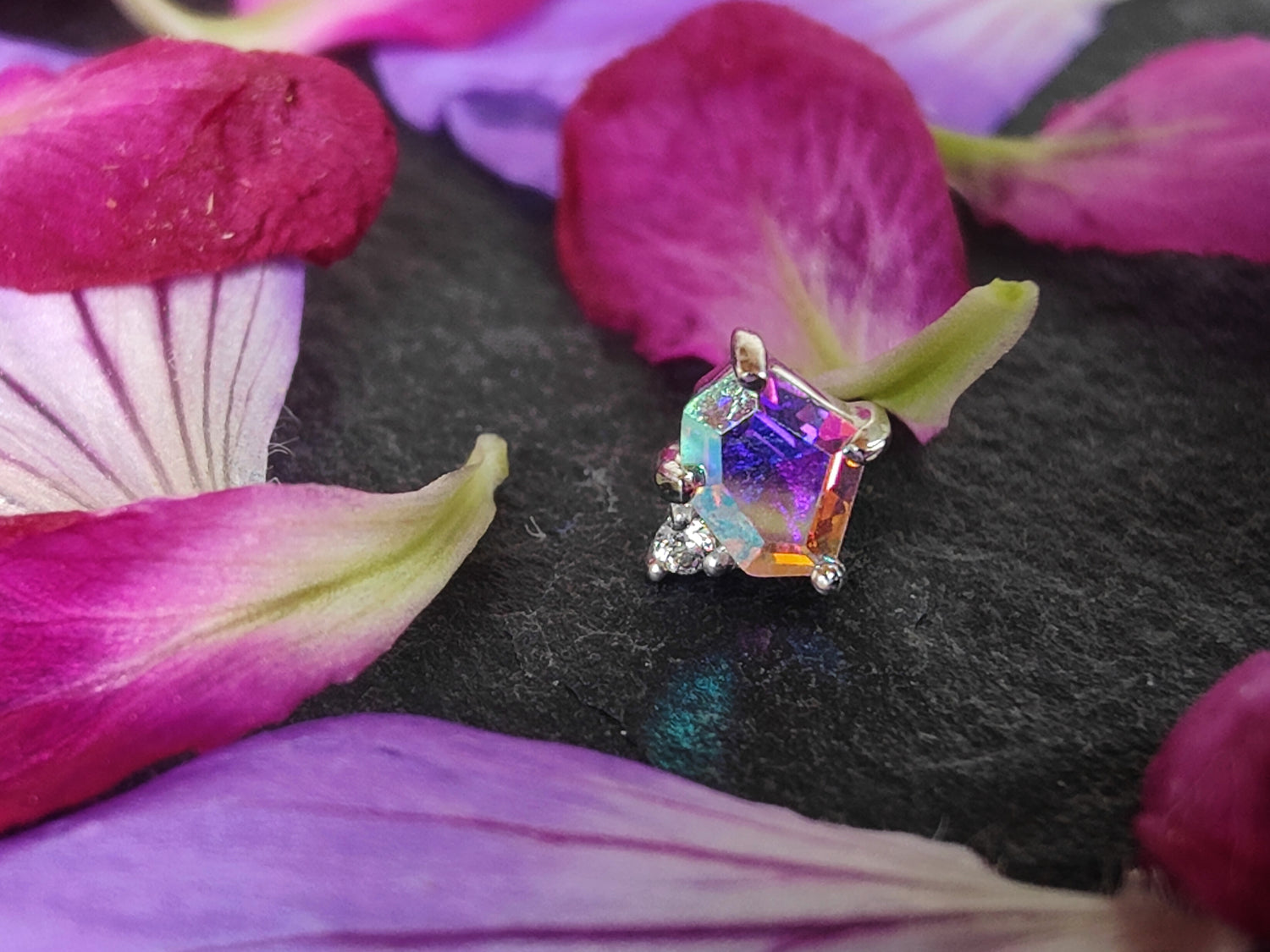 A photo of a piece of 14ct white gold body jewellery on a black piece of slate, surrounded by pink and purple flower petals. The piece of jewellery has one large asymmetrical hexagonal stone made out of mercury mist topaz, a stone which shines a rainbow colour, and connected to a much smaller white cubic zirconia. 