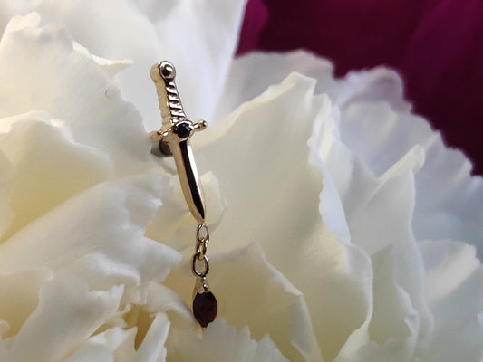 a yellow gold attatchement in the shape of a sword with a red garnet stone in the handle. connected is a charm of a marquise cut garnet stone. Sat on a white flower