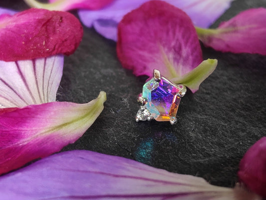 a asymmetrical hexgaon shaped stone in mercury mist topaz with a small white cz stone connected. Placed on a piece of slate surrounded by pink and purple petals
